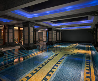 Indoor swimming pool with tiled mosaic floor, illuminated by blue recessed ceiling lights and surrounded by black pillars and walls. Steps lead into the water on both sides.