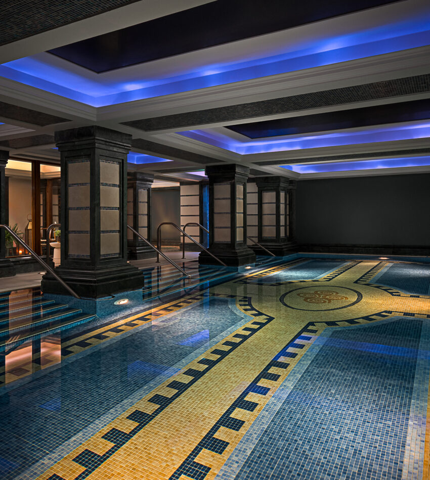 Indoor swimming pool with tiled mosaic floor, illuminated by blue recessed ceiling lights and surrounded by black pillars and walls. Steps lead into the water on both sides.