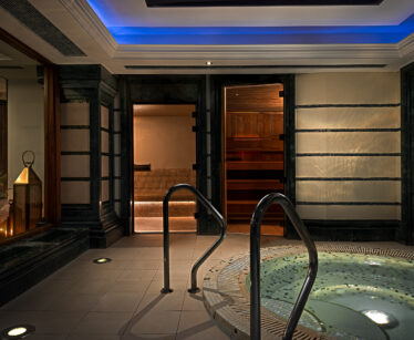 Indoor spa area featuring a round whirlpool hot tub with metal handrails, tiled floor, and a sauna room with wooden interior visible in the background.