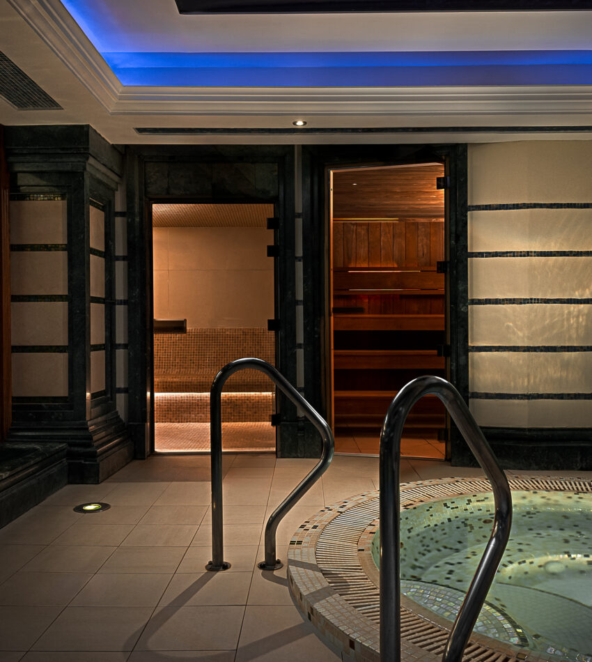 Indoor spa area featuring a round whirlpool hot tub with metal handrails, tiled floor, and a sauna room with wooden interior visible in the background.