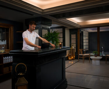 A man in a white polo shirt stands behind a dark reception desk, using a tablet in a modern, dimly lit lobby with plants and seating visible in the background.