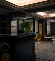 A dimly lit, modern hotel lobby with a black reception desk, potted plants, wooden chairs, and a display shelf with glassware and bottled water.