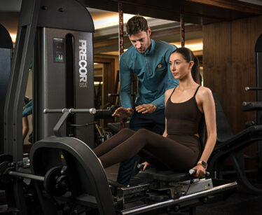 A woman uses a leg press machine at the gym whilst a male trainer stands behind her giving instructions.