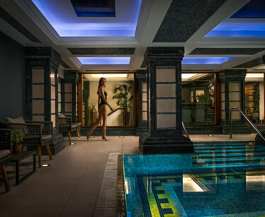 A woman in a black swimming costume walks along the edge of an indoor pool with blue lighting and decorative columns.