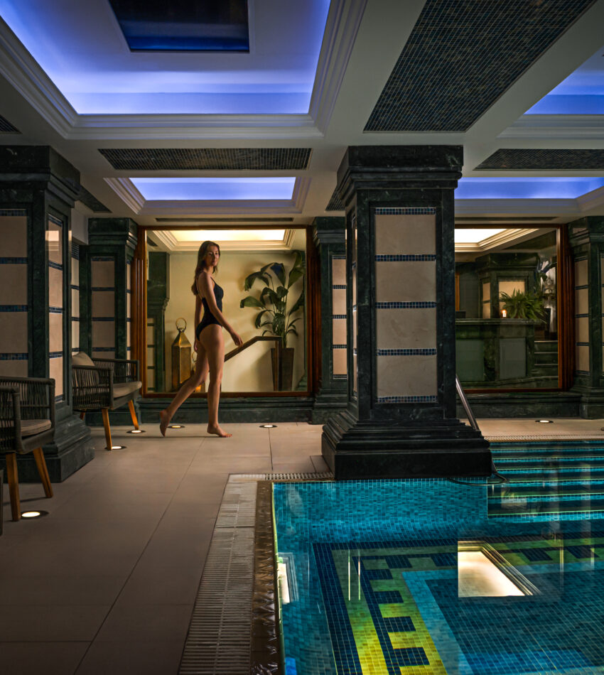 A woman in a black swimming costume walks along the edge of an indoor pool with blue lighting and decorative columns.