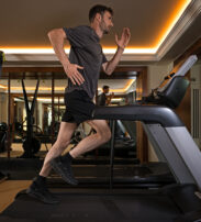 A man in sportswear runs on a treadmill in a modern gym with mirrored walls and exercise equipment visible in the background.