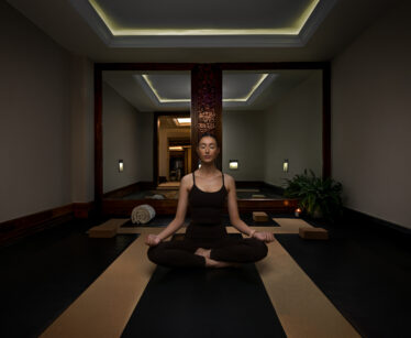A person sits cross-legged on a mat in a dimly lit room, meditating with eyes closed, surrounded by yoga props and a rolled towel.