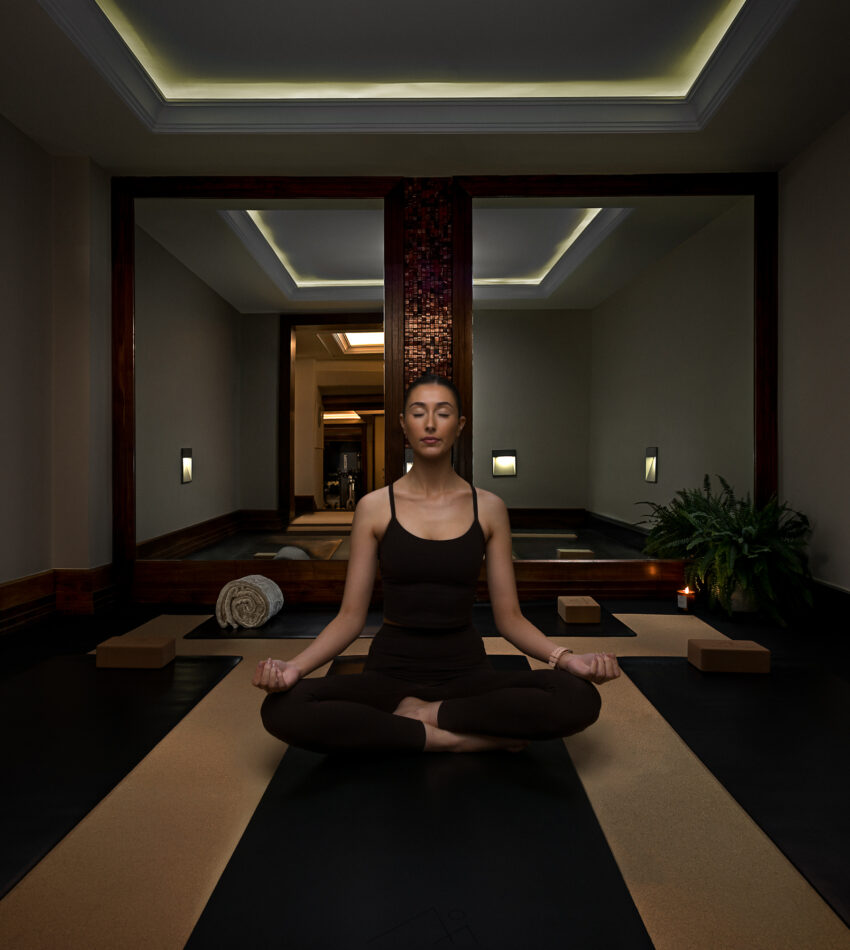 A person sits cross-legged on a mat in a dimly lit room, meditating with eyes closed, surrounded by yoga props and a rolled towel.