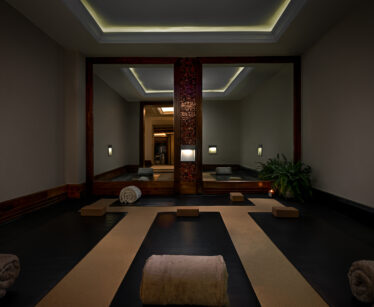 A dimly lit yoga or meditation room with large mirrors, soft lighting, mats, towels, blocks, and a plant in the corner.