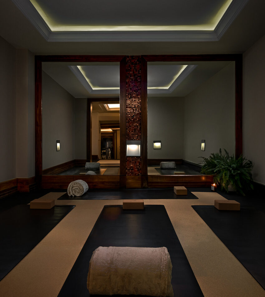 A dimly lit yoga or meditation room with large mirrors, soft lighting, mats, towels, blocks, and a plant in the corner.