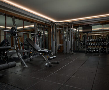 Modern gym room with weight benches, dumb-bells, barbells, cable machine, mirrored walls, and rubber flooring.