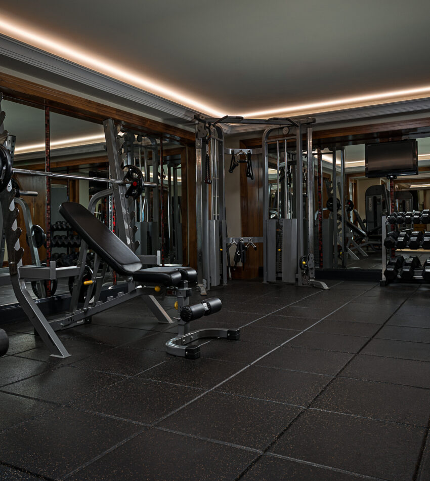 Modern gym room with weight benches, dumb-bells, barbells, cable machine, mirrored walls, and rubber flooring.