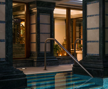 Indoor swimming pool with tiled columns and a stainless steel handrail, surrounded by glass doors and warm interior lighting.