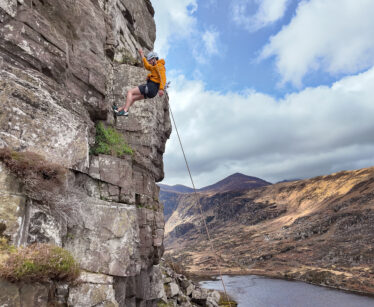 A person in an orange jacket and helmet climbs a rocky cliff with a safety rope, overlooking a river and hills under a partly cloudy sky.