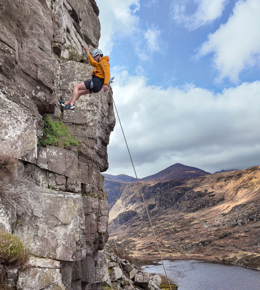 A person in an orange jacket and helmet climbs a rocky cliff with a safety rope, overlooking a river and hills under a partly cloudy sky.