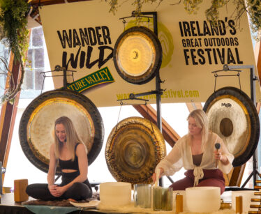 Two women on stage with gongs and sound bowls at the Wander Wild Ireland’s Great Outdoors Festival. One plays instruments; the other holds a microphone.