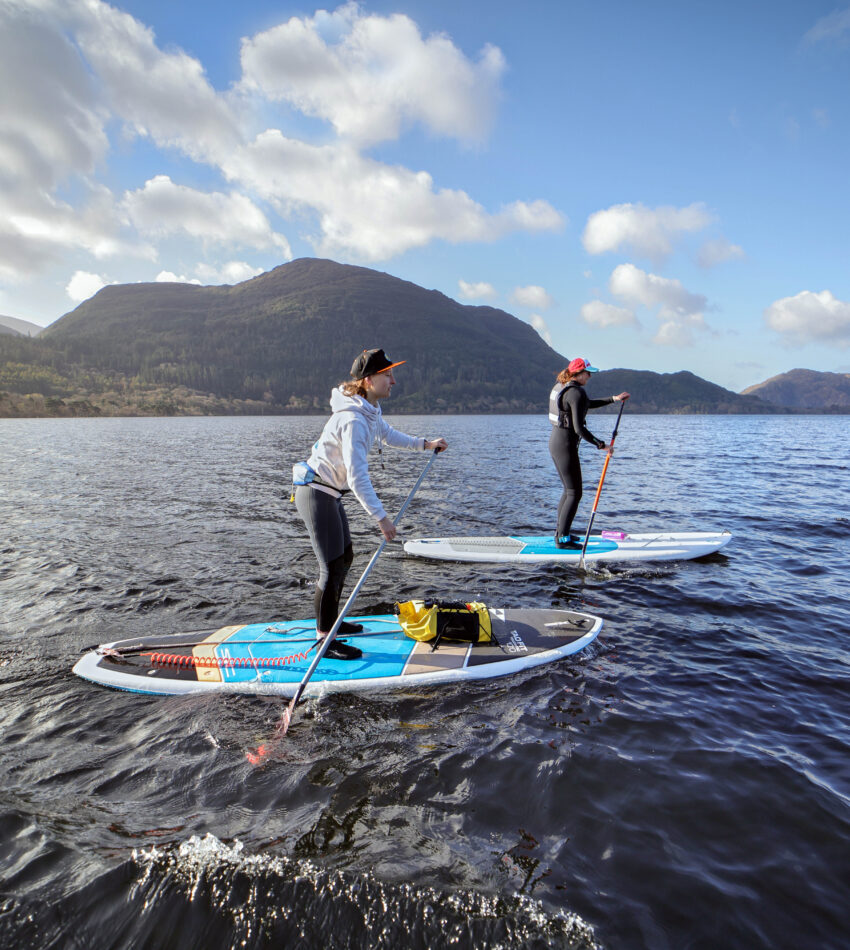 Two people stand-up paddleboarding on a lake, surrounded by mountains under a partly cloudy sky.