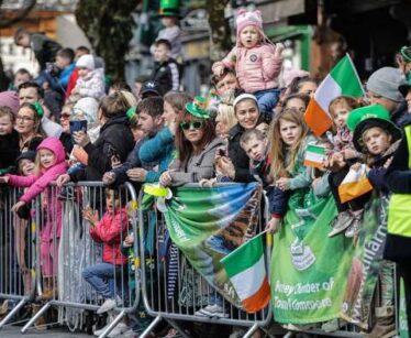 A crowd of people, many holding Irish flags and wearing green, stand behind barriers at an outdoor event or parade.