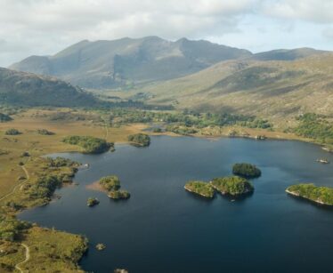 Aerial view of a lake with small islands, surrounded by green hills and mountains under a partly cloudy sky.