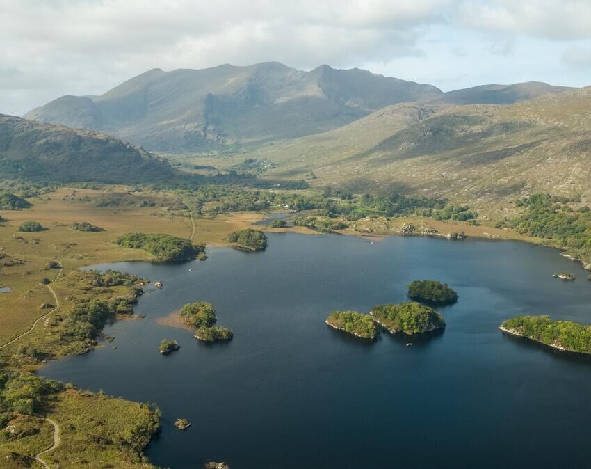 Aerial view of a lake with small islands, surrounded by green hills and mountains under a partly cloudy sky.