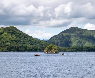 Small boats with people on a lake, with a small rocky island in the centre and green mountains in the background under a cloudy sky.