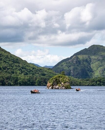 Small boats with people on a lake, with a small rocky island in the centre and green mountains in the background under a cloudy sky.