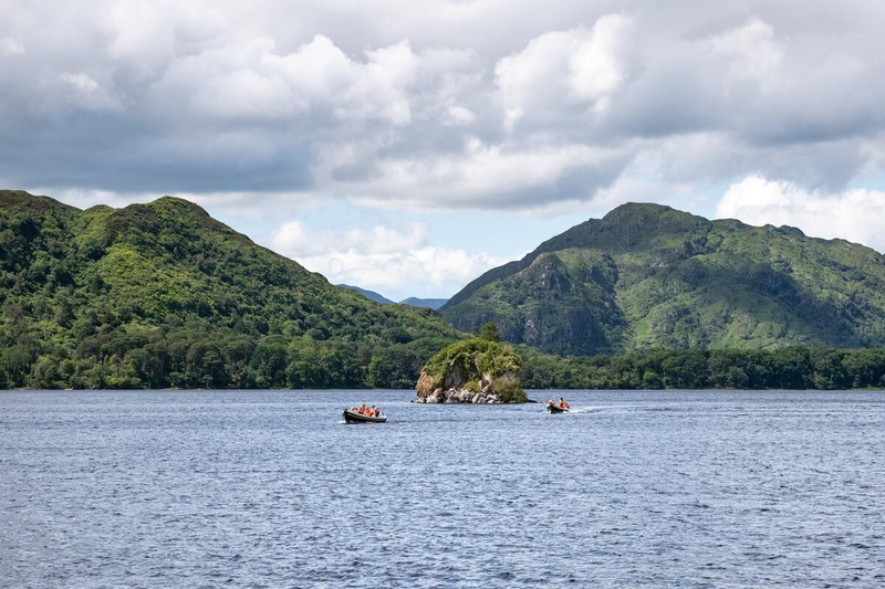 Small boats with people on a lake, with a small rocky island in the centre and green mountains in the background under a cloudy sky.