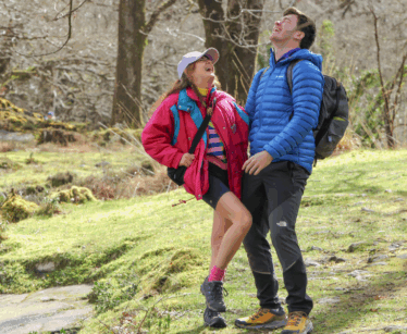 Two people dressed in outdoor clothing and rucksacks stand on a forest trail, smiling and laughing together on a sunny day.