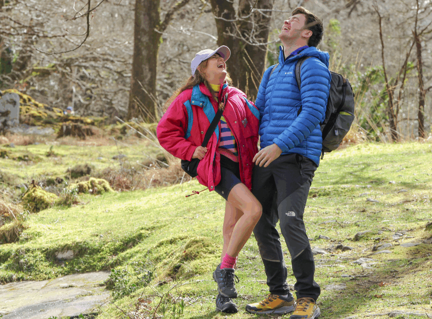 Two people dressed in outdoor clothing and rucksacks stand on a forest trail, smiling and laughing together on a sunny day.