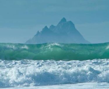 Waves crash in the foreground with a large, jagged mountain rising in the distance under a cloudy sky.
