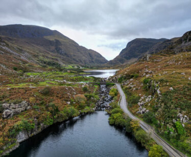 A winding road runs beside a river and valley, surrounded by rocky hills and mountains under a cloudy sky.