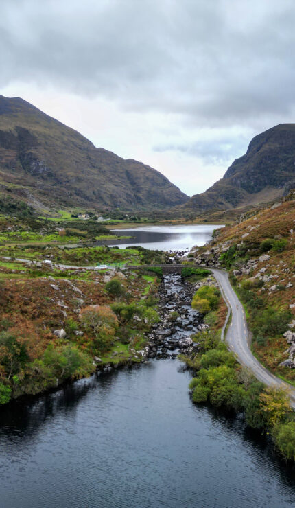 A winding road runs beside a river and valley, surrounded by rocky hills and mountains under a cloudy sky.