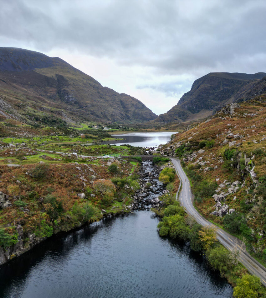 A winding road runs beside a river and valley, surrounded by rocky hills and mountains under a cloudy sky.