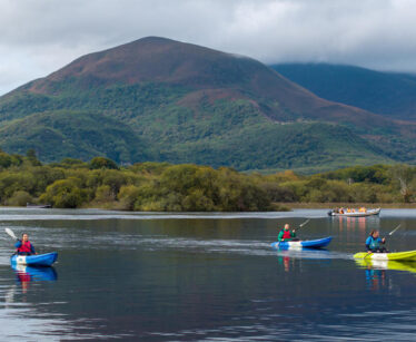 Three people kayak on a calm lake with green hills and a large mountain in the background under a cloudy sky.