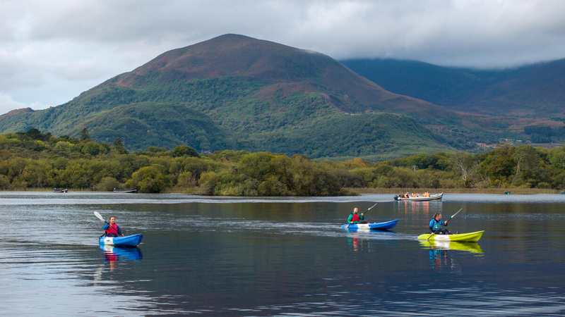 Three people kayak on a calm lake with green hills and a large mountain in the background under a cloudy sky.