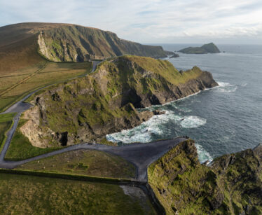Aerial view of a rugged coastline with steep cliffs, winding roads, and waves crashing against the rocky shore under a partly cloudy sky.