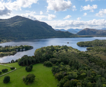 Aerial view of a large lake surrounded by green fields, dense forests, and distant mountains under a partly cloudy sky.
