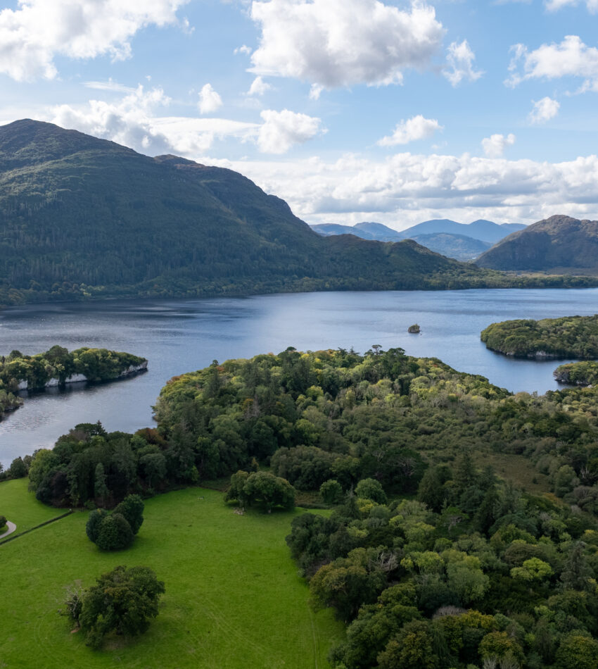 Aerial view of a large lake surrounded by green fields, dense forests, and distant mountains under a partly cloudy sky.