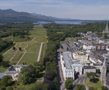 Aerial view of a town with a park on the left, buildings on the right, and mountains and a lake in the background under a clear sky.
