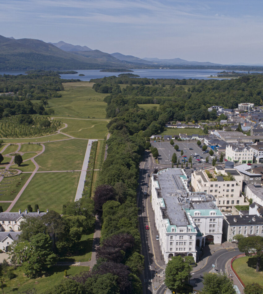 Aerial view of a town with a park on the left, buildings on the right, and mountains and a lake in the background under a clear sky.