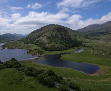 Aerial view of a lush green landscape with a hill, small lakes, and distant mountains under a partly cloudy sky.
