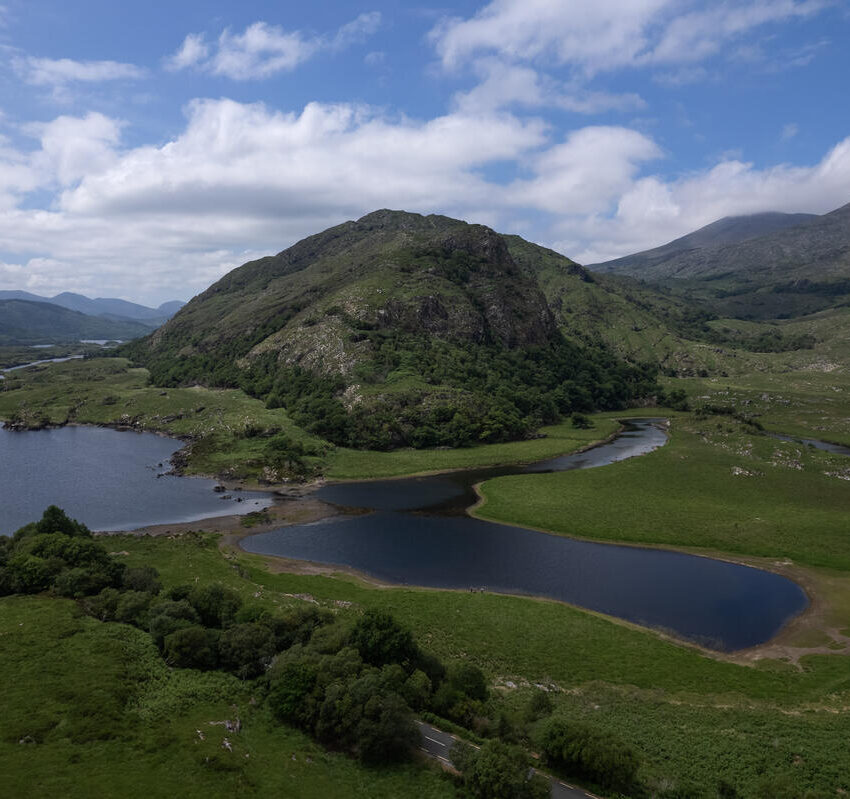 Aerial view of a lush green landscape with a hill, small lakes, and distant mountains under a partly cloudy sky.