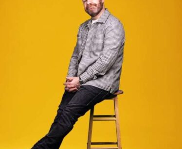 Man wearing a checked shirt and jeans sits on a wooden stool against a solid yellow background.