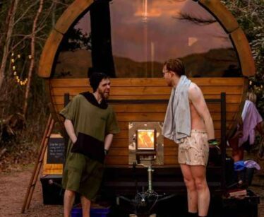Two men stand talking outside a small, round wooden sauna with a glass door, surrounded by trees. One man is barefoot and the other wears sandals and has a towel over his shoulder.