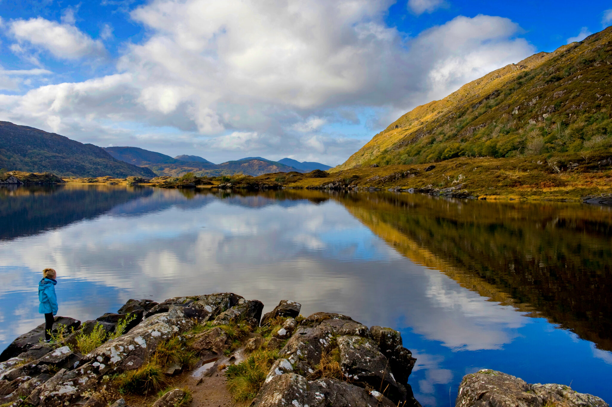A person in a blue jacket stands on rocky terrain by a calm lake with mountains and clouds reflected in the water under a partly cloudy sky.