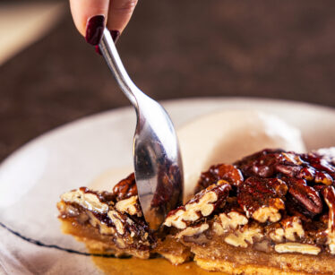 Close-up of a hand with dark nails enjoying pecan pie with vanilla ice cream at a hotel restaurant, on a marble table.