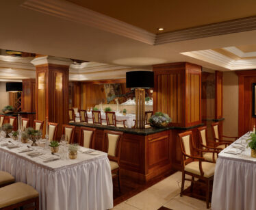 A formal dining room with two long tables set with white tablecloths, candles, and greenery, surrounded by wooden columns and cream-coloured chairs.