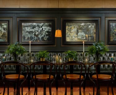 A long dining table with wooden chairs, green plants, lit candles, and dark tufted seating beneath three abstract paintings on a dark-panelled wall.