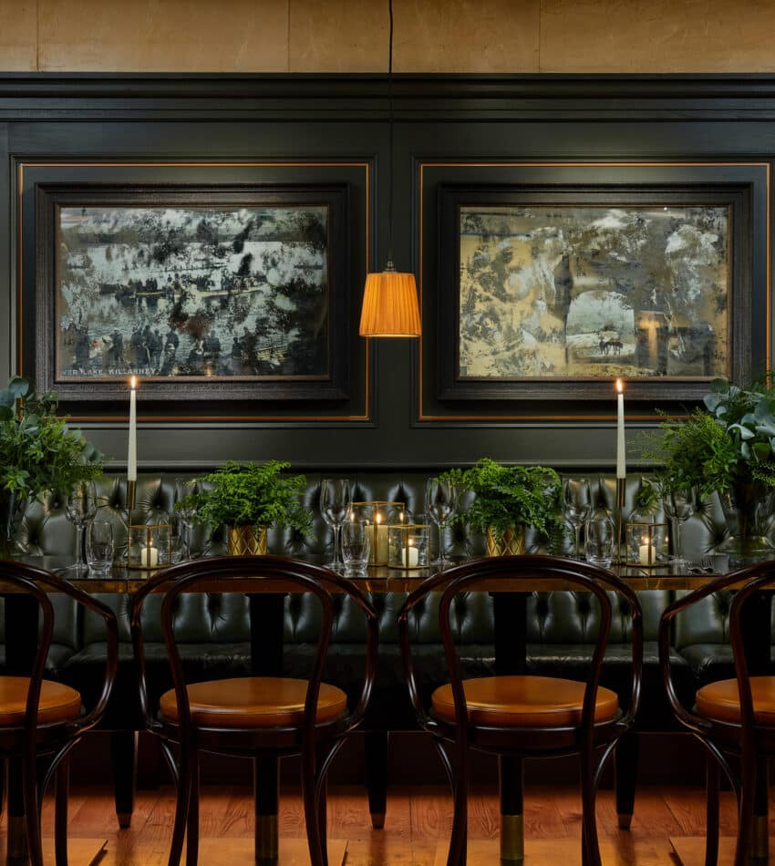 A long dining table with wooden chairs, green plants, lit candles, and dark tufted seating beneath three abstract paintings on a dark-panelled wall.