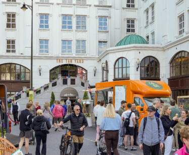 People gather around food vans and stalls outside a white building called "The Tan Yard" during a street event. Some people are walking, eating, or cycling.